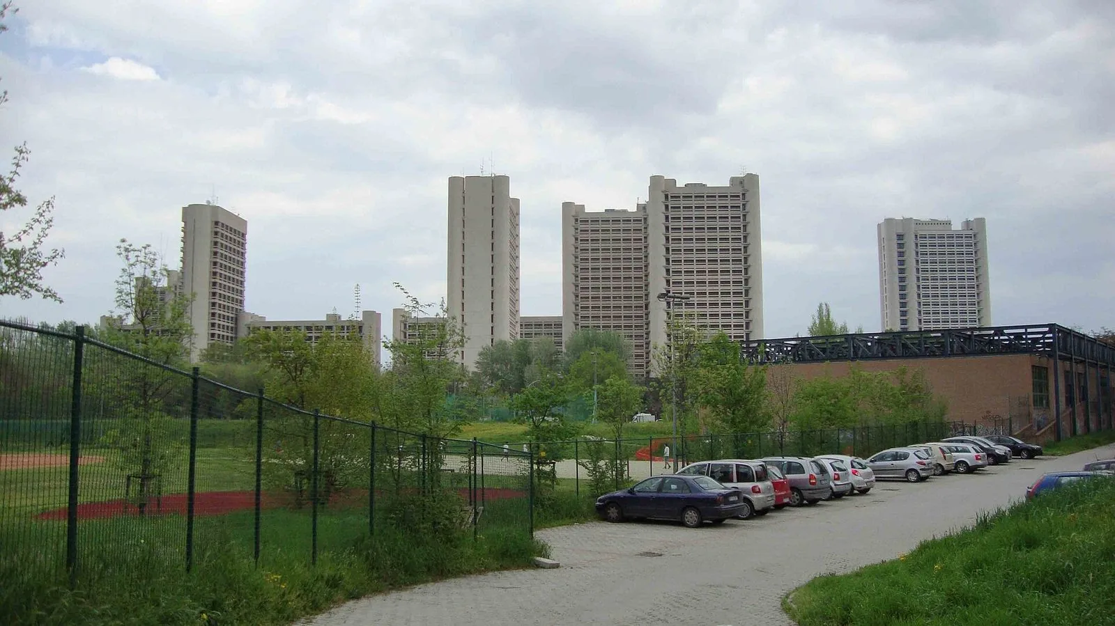 Le torri chiare in cemento del Fiera District di Bologna, progettato da Kenzo Tange, viste da un parco con alberi e auto parcheggiate sotto un cielo nuvoloso