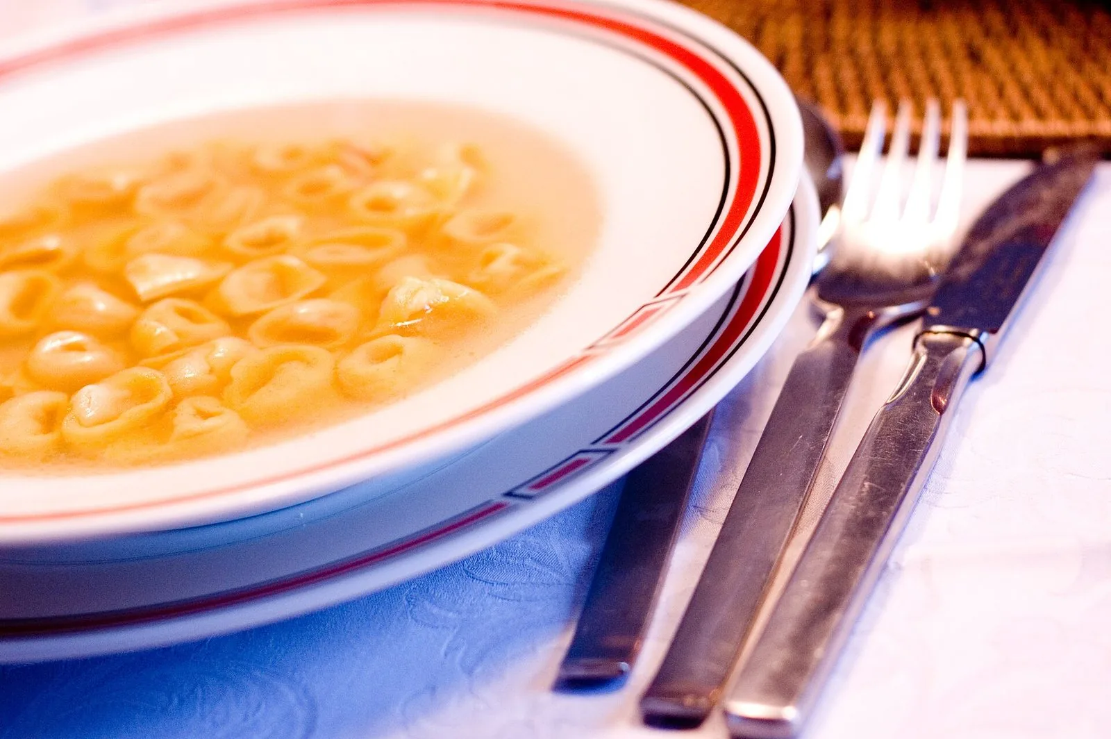Un piatto fondo bianco con il bordo rosso pieno di tortellini in brodo dorato, accanto a forchetta, coltello e cucchiaio su una tovaglia chiara