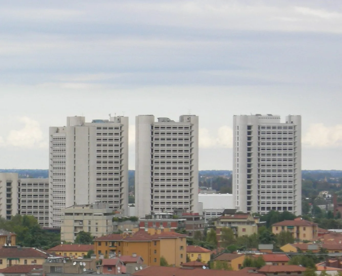 Tre alte torri bianche per uffici del Fiera District di Bologna viste dall'alto, oltre i tetti rossi e le facciate gialle di case e palazzine, sotto un cielo grigio