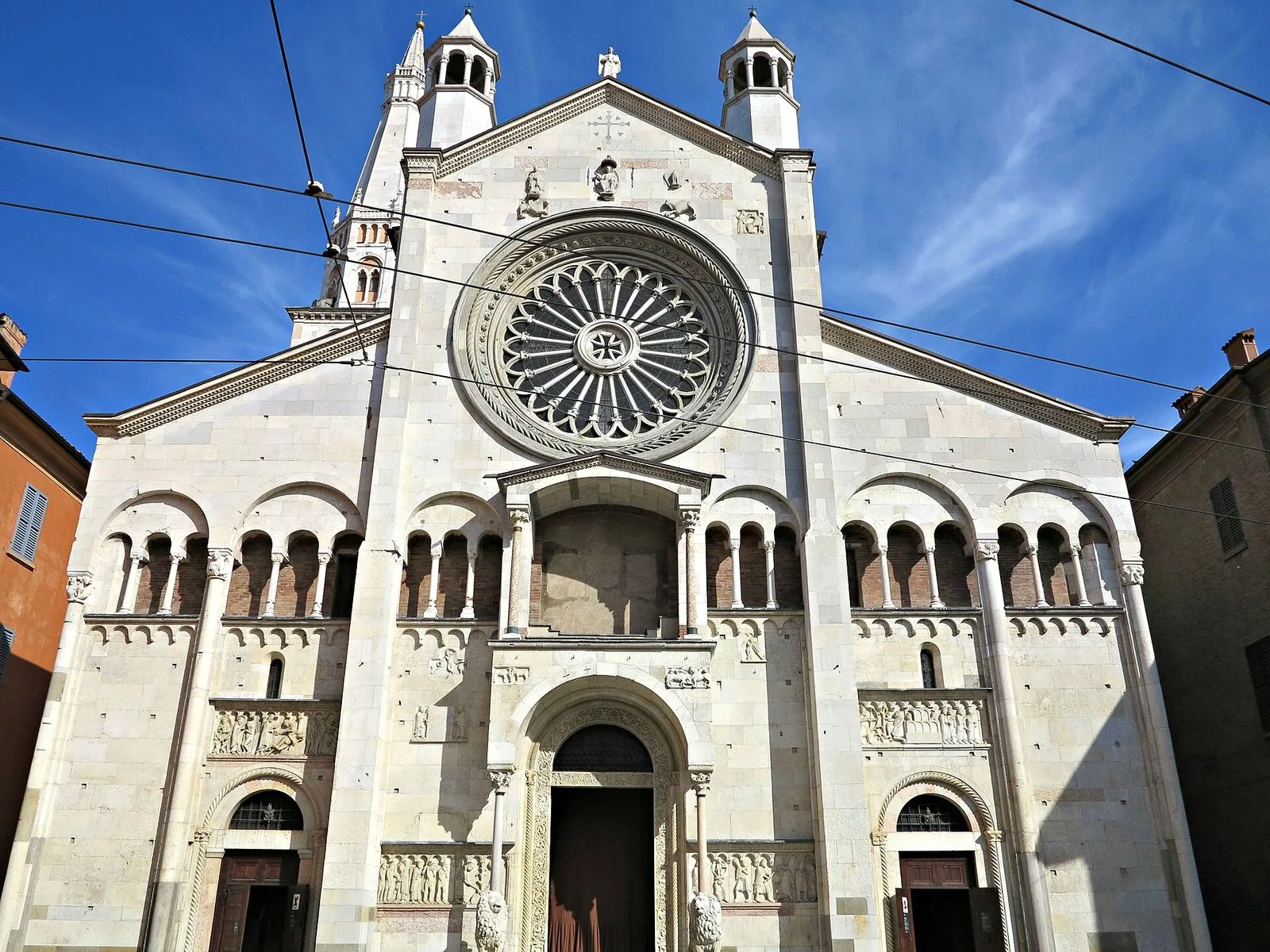 La facciata in pietra chiara del Duomo di Modena sotto un cielo azzurro, con il grande rosone, la loggia ad archetti, i rilievi scolpiti e il portale centrale con i leoni, e a sinistra la cima della torre Ghirlandina