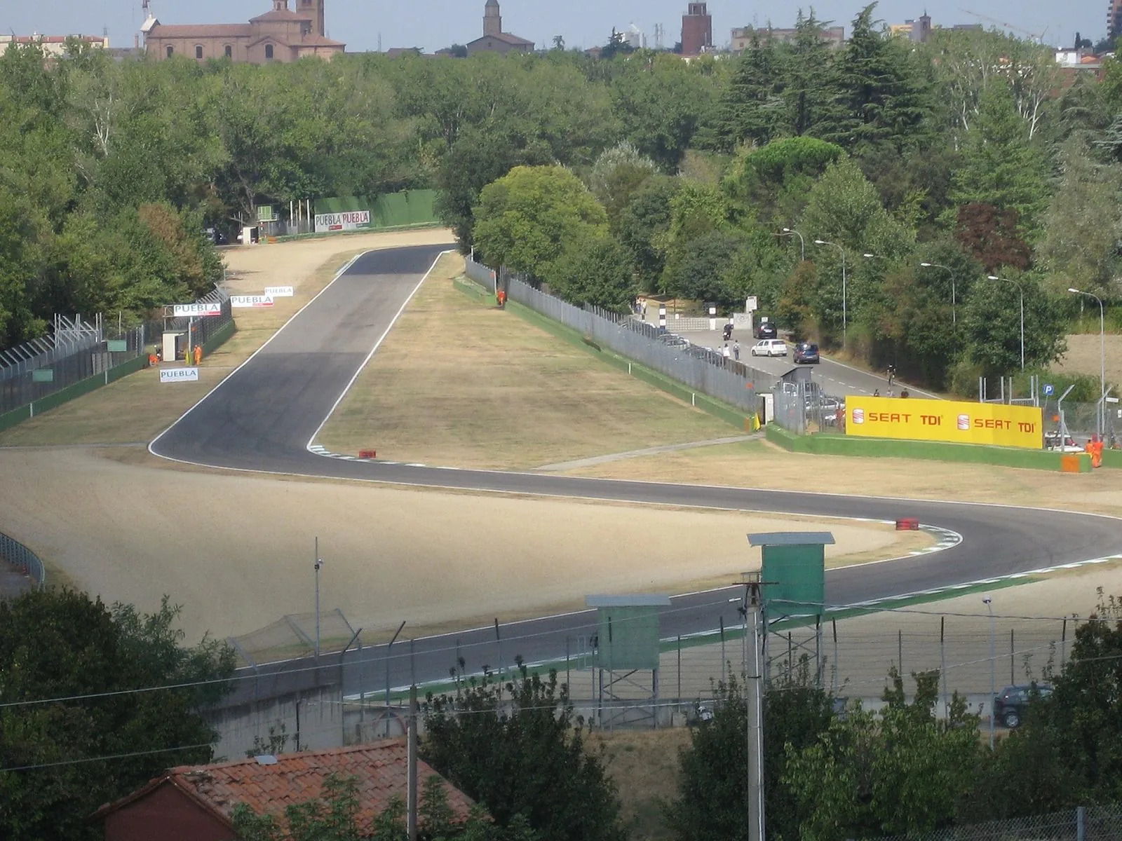 Il circuito di Imola visto dall'alto: la pista d'asfalto disegna una curva a esse tra vie di fuga in ghiaia e prati secchi, circondata da alberi, con i campanili e i tetti della città sullo sfondo