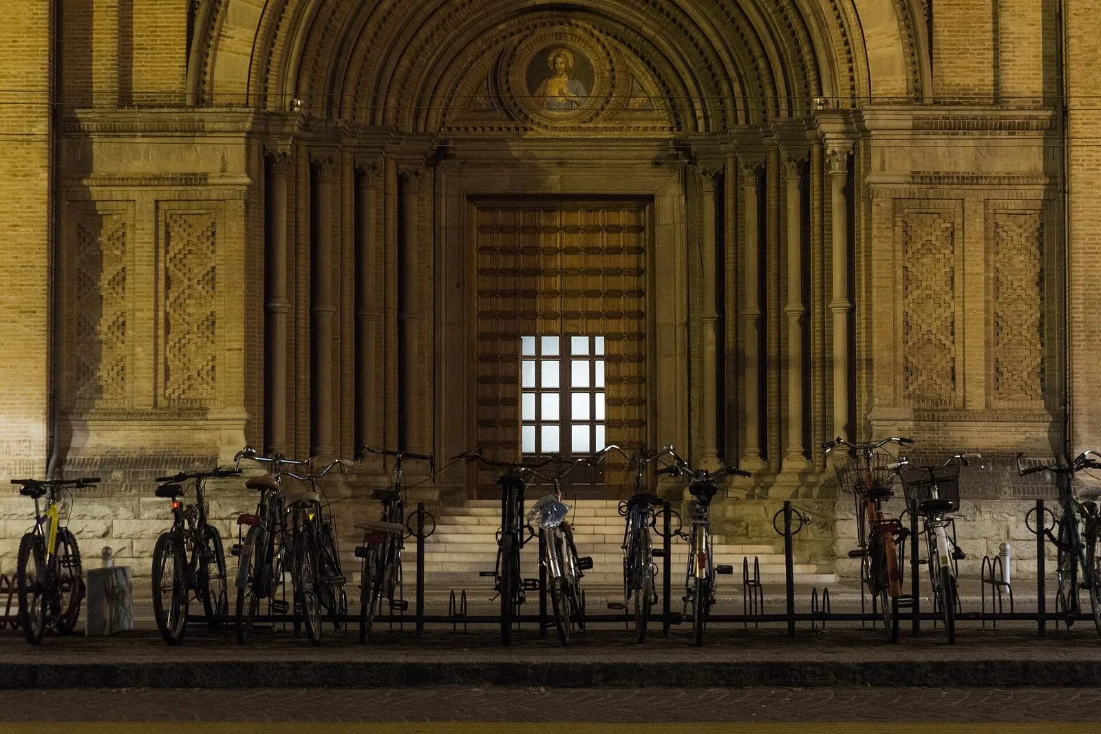 Una fila di biciclette parcheggiate nelle rastrelliere davanti al grande portale in mattoni di una chiesa di Bologna, illuminato di sera, con un mosaico nella lunetta