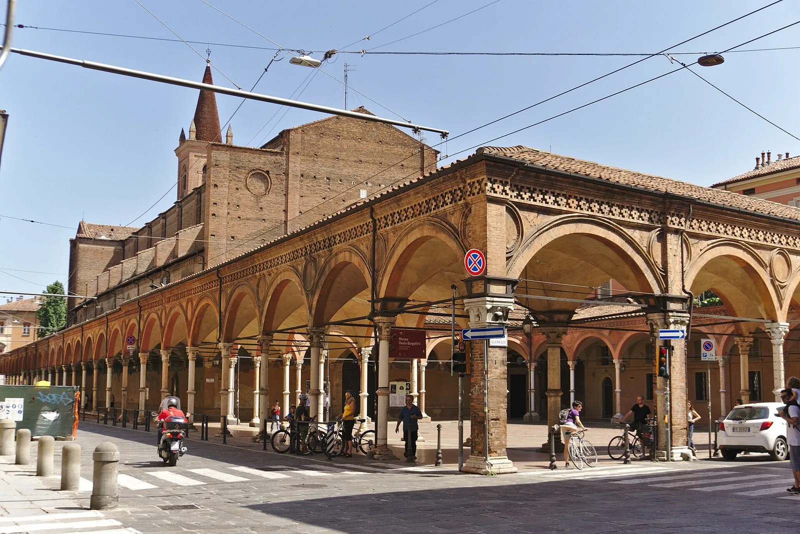 Il portico di Santa Maria dei Servi su Strada Maggiore, con le arcate su colonne sottili e la decorazione in cotto, la fiancata in mattoni della basilica e il campanile appuntito in una giornata di sole, con passanti e biciclette all'angolo della strada